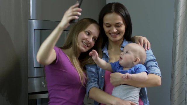 Medium shot of happy lesbian couple with baby boy smiling and taking selfie on mobile phone