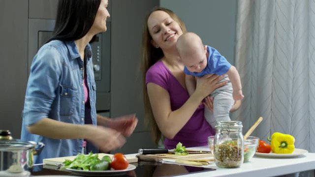 Tilt up of happy young woman cutting vegetables in kitchen, then taking cute baby boy from her female partner and kissing him on cheek