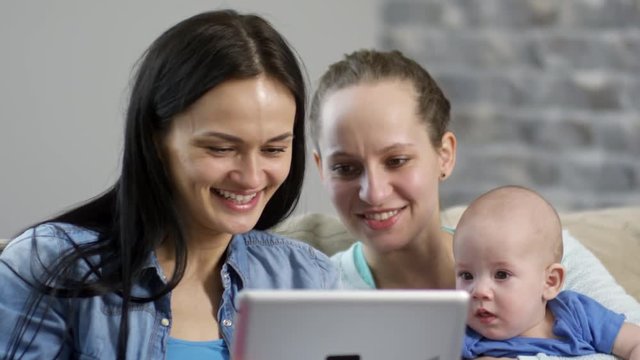 Handheld medium shot of happy female couple with baby boy watching cartoons on tablet and laughing