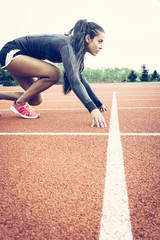 Athlete crouched in starting position on a running track. Ethnic, multicultural theme. Wearing long sleeve black top, shorts and pink runners. Long black hair in a pony tail.