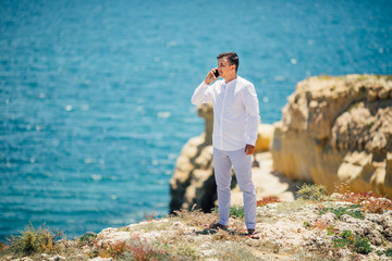Stylish young man man talking on phone standing on the cliff along the ocean beach
