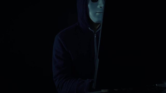 Dangerous masked hacker using a computer for cyber attack, phishing and hacking security system in absolutely dark environment with copy space (using panning technique)