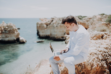 Senior man working on his laptop sitting on the top of cliff on the beach