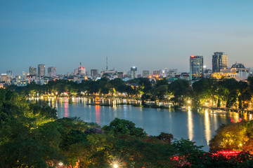 Fototapeta premium Hoan Kiem lake lub Sword lake, Ho Guom w Hanoi, Wietnam z Turtle Tower, zielone drzewa i budynki na horyzoncie, o zmierzchu