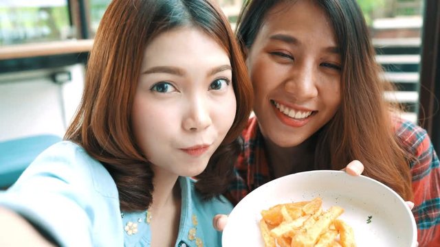 Beautiful happy Asian women lesbian lgbt couple sitting each side eating a plate of Italian seafood spaghetti and french Fries at restaurant or cafe. Female couple using smartphone for selfie.