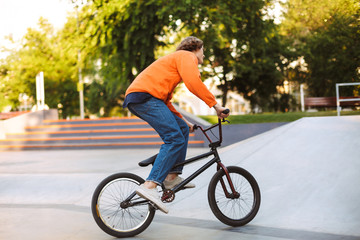 Young guy in orange pullover and jeans from back riding bicycle at skatepark