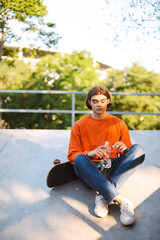 Young guy in orange pullover with skateboard holding bottle of water in hand while spending time at skatepark