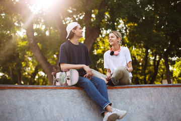 Smiling girl with headphones and young guy with skateboard joyfully spending time together at...