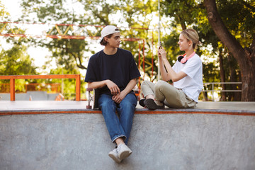 Naklejka na ściany i meble Smiling girl with headphones taking photo of young guy with skateboard on cellphone spending time together at modern skatepark