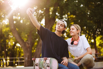 Smiling girl with headphones and young guy with skateboard happily taking photos on frontal camera...