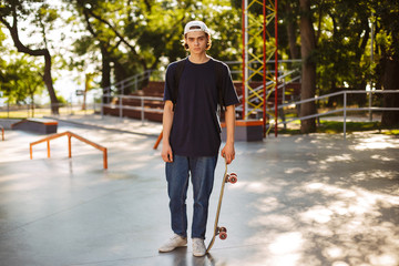 Young skater in white cap dreamily looking in camera holding skateboard in hand with modern skatepark on background