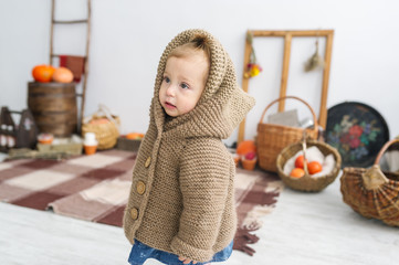 Cute little girl in warm clothes standing in white big room with pumpkins.