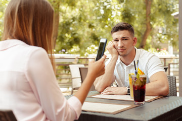 Young couple drinking coffee in a cafe. Serious man worriedly looking while his girl using smart phone
