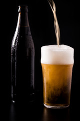 Pouring crafted unfiltered beer with foam in glass with bottle behind isolated on black dark background with soft reflections and beautiful shadows. Studio shot.