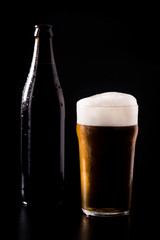 Glass of crafted unfiltered beer with foam and bottle behind isolated on black dark background with soft reflections and beautiful shadows. Studio shot.