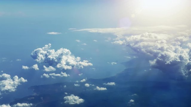 Flying along the sea coast through fluffy clouds in pure sunshine. Picturesque view from airplane window to rural coastline with sun shining brightly over the clouds with beautiful rays and lens flare