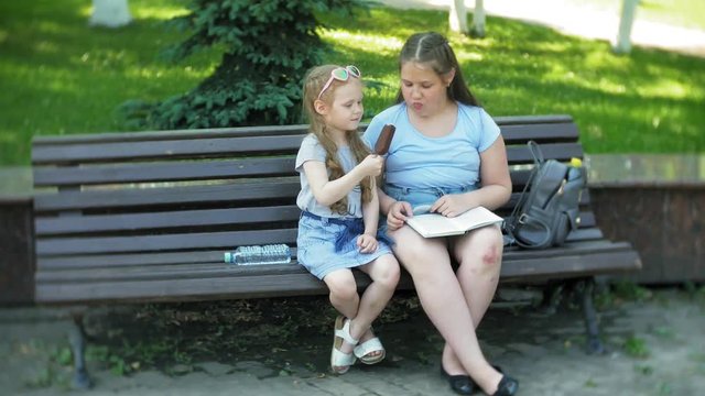 Two little girls are sitting on a wooden bench in a city reading a book and eating ice cream, the background of a city park