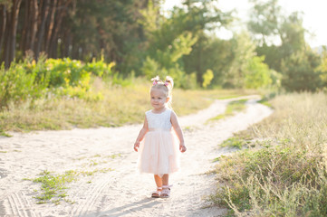 Healthy, lifestyle, happy childhood concept. Adorable little child. Girl with blonde hair, wearing a pink dress. A girl walks with flowers in nature on forest road of the setting sun. Children