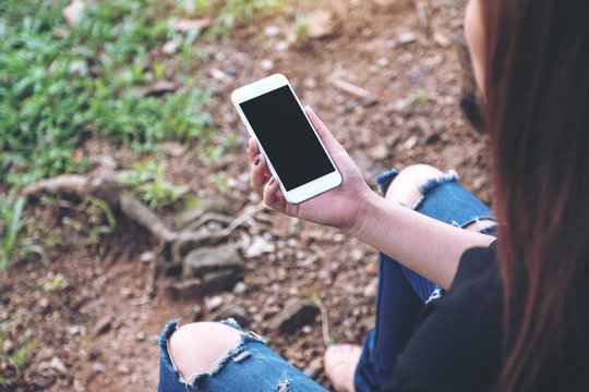Mockup image of woman holding white mobile phone with blank black desktop screen while sitting on the ground outdoor