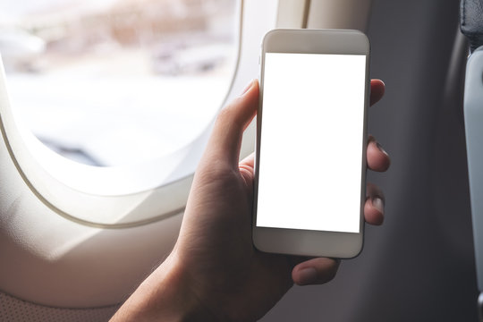 Mockup image of a hand holding a white smart phone with blank desktop screen next to an airplane window