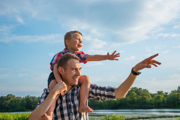 Happy young father holds his son piggyback ride on his shoulders, points at something with his hand...