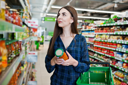 Shopping woman looking at the shelves in the supermarket.  Portrait of a young girl in a market store holding green shop basket and can of vegetables.