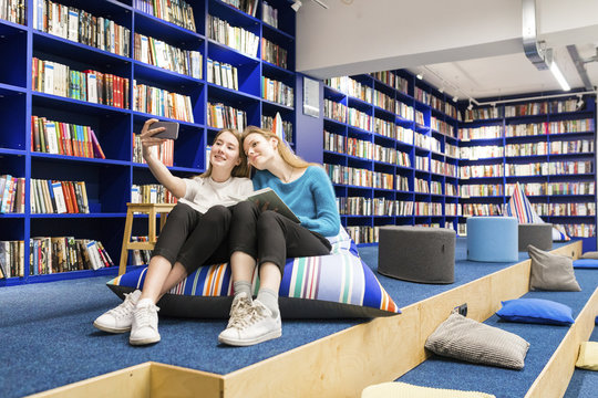Two teenage girls sitting on beanbag in a public library taking selfie with smartphone