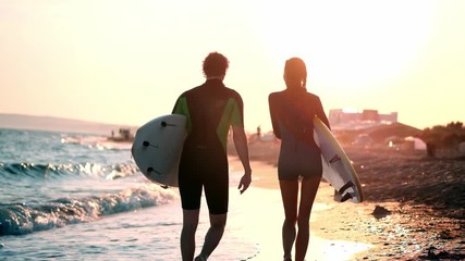 SLOW MOTION: A young couple of guy and girl surfers are walking along the beach in wetsuits. In the hands of holding surfboards. Look at the waves and the sunset - Powered by Adobe