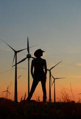 A young girl stands in a field and admires the wind turbines  in the distance. The sun is setting and the sunlight creates a  silhouette to the scenery.  