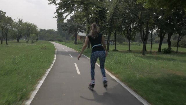 White handsom brown hair girl in tight blue pants woman riding roller skates on bike road in park, during summer day