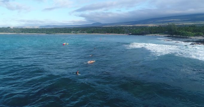Slow motion aeril footage of a girl in bikini surfing a wave with a long beach and a conoe in the background in Kailua Kona, Hawaii