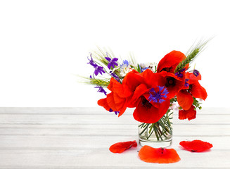 Naklejka premium Red poppies, cornflowers, ears of green wheat, chamomile in small vase on white wooden table on white background with space for text.