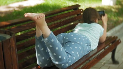 Little fat girl with a tablet PC and headphones sitting on a bench listening to music or watching a video in a summer park