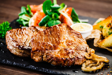 Grilled steaks with baked potatoes and vegetables served on black stone on wooden table 