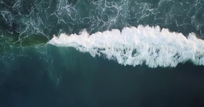 Aerial top view waves break on dark rocks near beach. Sea waves on the dangerous stones aerial view drone 4k shot. Bird's eye view of ocean waves crashing against an empty beach from above.