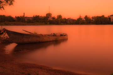The boat near the shore of the river or the lake in the rays of setting or rising sun. The summer countryside landscape in the warm orange colours