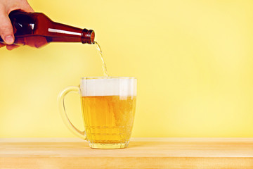A man pours beer into a mug from a bottle on a yellow background on a wooden table. Copy space.