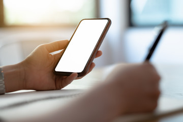 Mockup image of a woman holding mobile phone and writing notebook with blank black screen in modern loft cafe