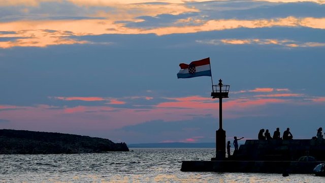 Stone pier with lighthouse and flying Croatian flag in sunset slow motion, people walking and sitting on stone wall, silhouettes of people only, individuals not recognizable