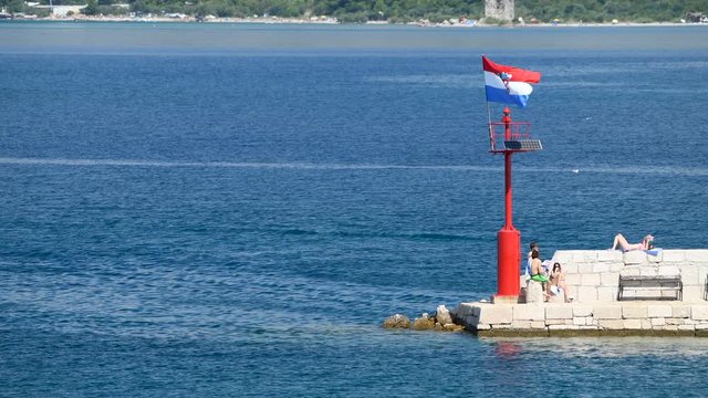 Stone pier with lighthouse and flying Croatian flag in daylight, people walking and sitting on stone wall