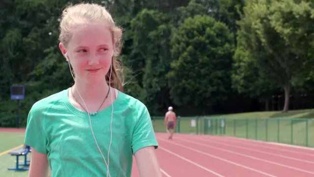 Camera preceding tilts up at Teen girl athlete walking on track and smirking at someone off camera in slow motion