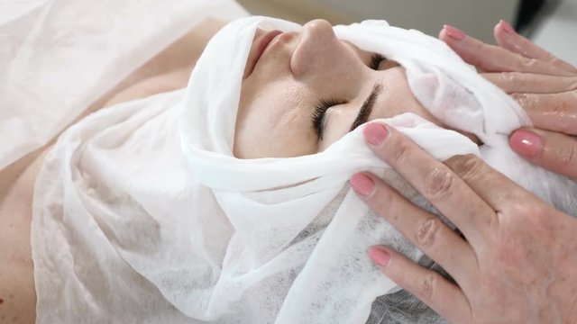 Beauty concept. Young female client in healthcare clinic. Woman gets procedure on face. Cosmetologist wraps face with towel to clean it