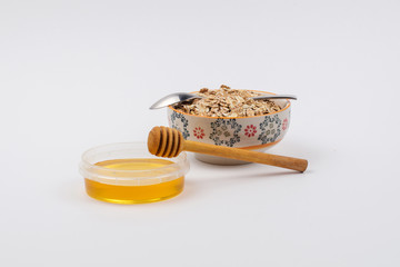 A ceramic bowl full of oat flakes and a container with honey, a stainless steel spoon and honey dipper on a white background