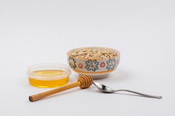 A ceramic bowl full of oat flakes and a container with honey, a stainless steel spoon and honey dipper on a white background