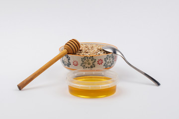 A ceramic bowl full of oat flakes and a container with honey, a stainless steel spoon and honey dipper on a white background