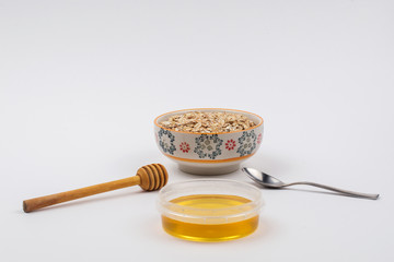 A ceramic bowl full of oat flakes and a container with honey, a stainless steel spoon and honey dipper on a white background