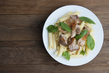 Cooked penne pasta isolated in white round plate, with basil leaves and pork meat, over wooden table, decorated with basil leaves. Top view.