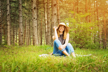 Young woman in summer boho style outfit, sitting in forest or park, smiling, enjoying nature. Female person in forest sitting on grass, posing. Natural lighting, retouched.