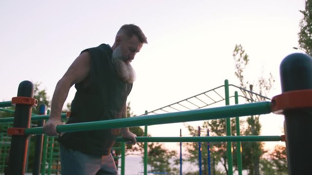 Fit handsome middle aged man with long gray beard doing push ups on parallel bars on sports ground with sea background