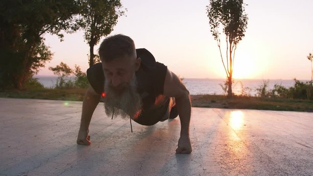 Fit handsome middle aged man with long gray beard doing push ups on sports ground with sea background